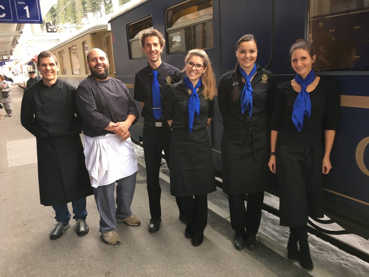 Die sechsköpfige RailGastro-Crew auf dem Bahnsteig, in Schwarz mit blauen Halstüchern, dahinter der königsblaue Speisewagen
