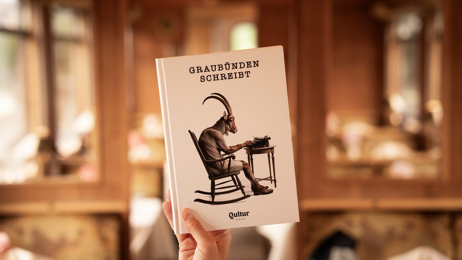 Eine Hand hält das Buch «Graubünden schreibt» hoch: auf dem Umschlag ein Steinbock, der im Schaukelstuhl an einer Schreibmaschine sitzt. Dahinter unscharf der holzgetäfelte Speisewagen