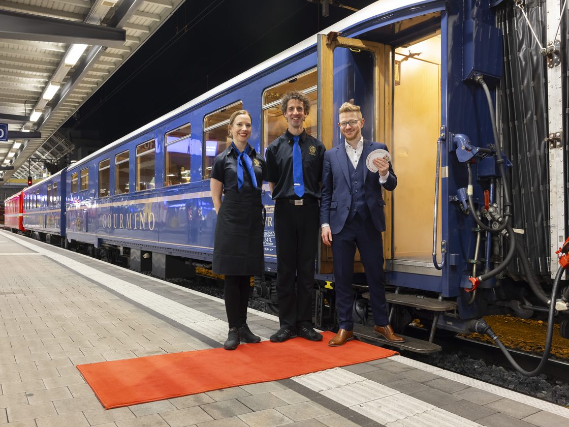 Drei Mitarbeitende auf einem roten Teppich vor dem blau-goldenen Speisewagen am Bahnsteig