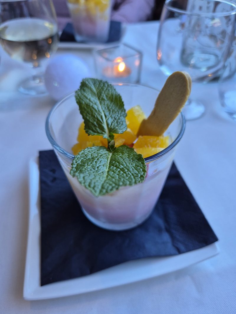 Ein Dessert im Glas mit Minzblatt auf einer Schieferplatte, dahinter ein gedeckter Tisch bei Kerzenlicht